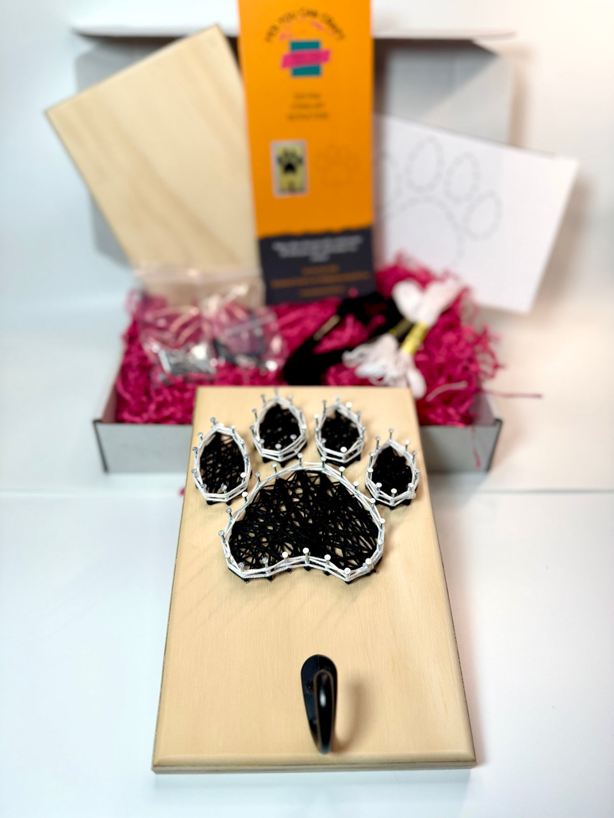 dog paw string art with hook for leash with kit contents in background including board, instructions, thread, hook, nails and mounting hardware.
