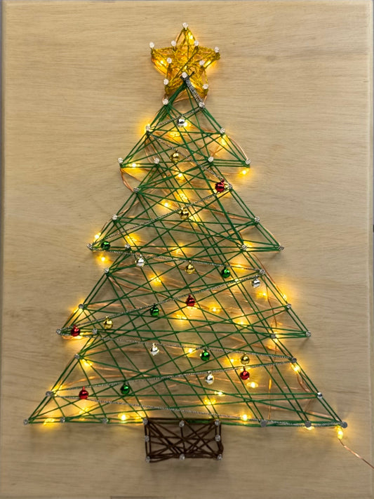 A string-art Christmas tree decoration on a light wood background, with green wrapped lines forming a tree shape, warm white string lights, small multicolored ornaments, a gold star topper, and a brown trunk base.
