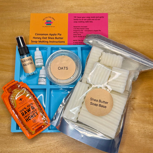 A DIY soap making kit laid out on a wooden surface, including a blue mold tray, a large bag of Shea Butter Soap Base, a round tin of OATS, a bottle of Rubbing Alcohol, two small dropper bottles, an orange packet of raw honey, and printed making instructions and flavor cards.