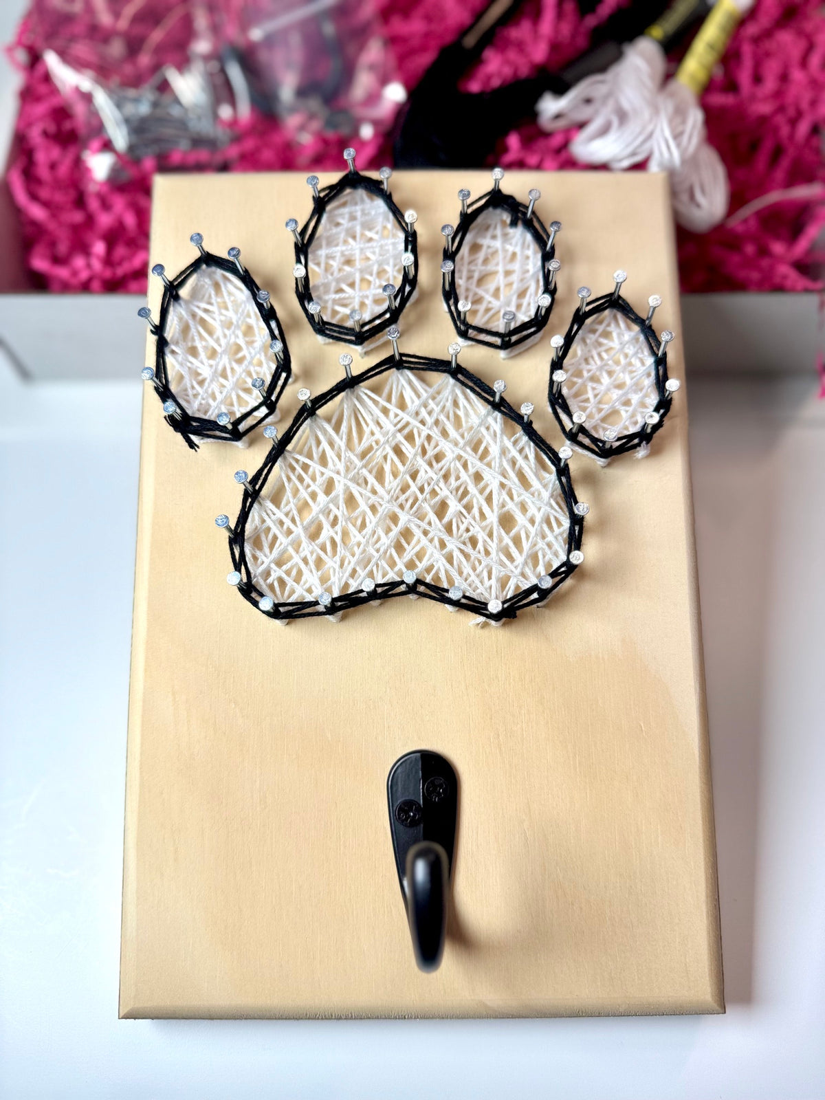 Dog paw string art piece with hook for hanging leash. White dog paw with black border. Contents of the kit in the background. 