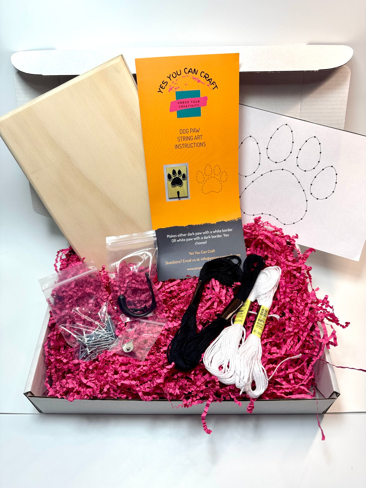 A DIY craft kit in an open box filled with bright pink paper shred, including a wooden block, printed instruction card, embroidery floss, black and white threads, metal findings, and a paw print template.
