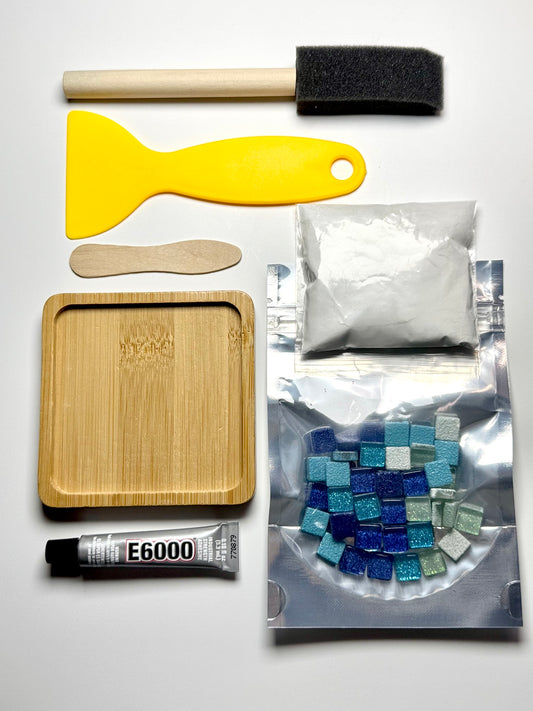 Mosaic tile kit components laid out on a white background, including a yellow spatula, wooden tools, a square bamboo coaster, a tube of E6000 adhesive, a bag of grout, and a bag of mosaic tiles.