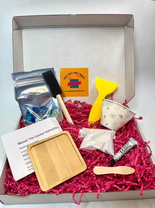A masonry mosaic tile coaster kit displayed inside its open cardboard box filled with bright pink shredded paper. The kit includes a small wooden tray, a white patterned cup, a yellow spatula, a black-handled brush, a bag of blue mosaic pieces, a tube of glue, instructions, and a small wooden tool, all arranged neatly.