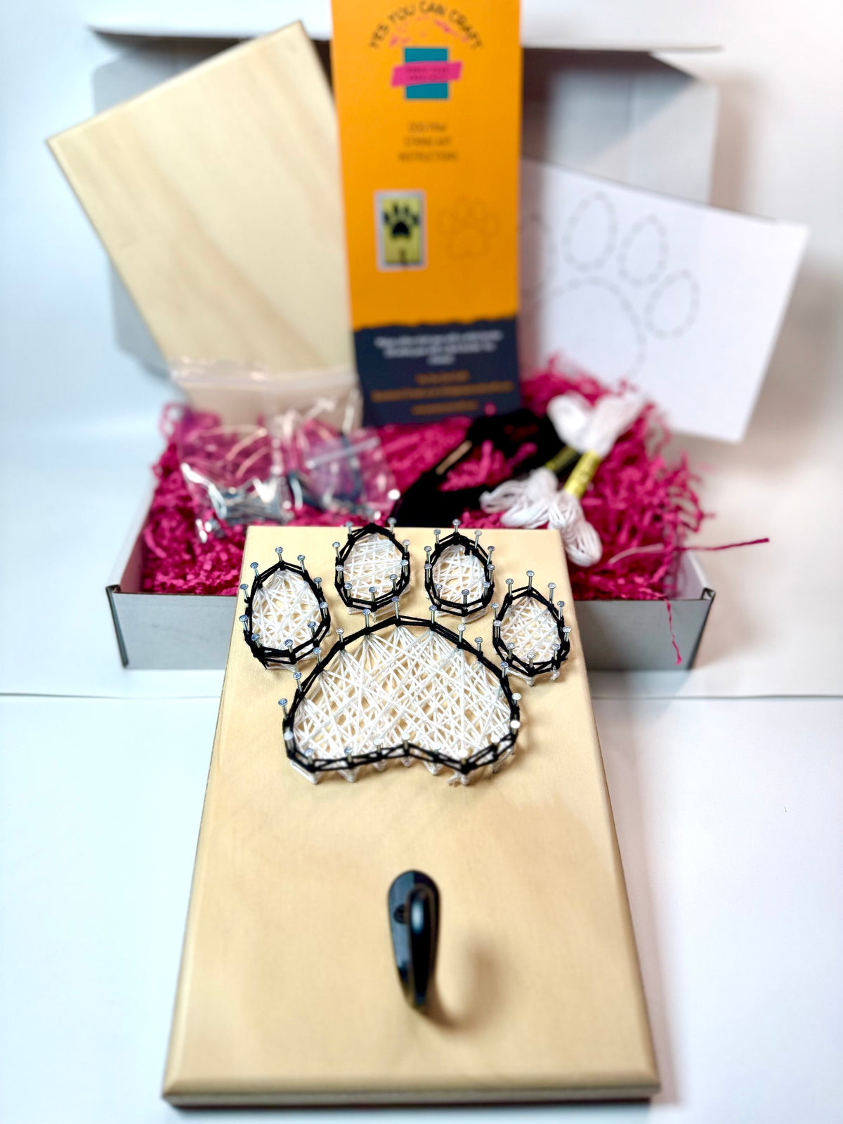dog paw string art piece with hook for hanging leash. contents of kit in the background include black and white thread, hook, nails, board, instructions and mounting hardware