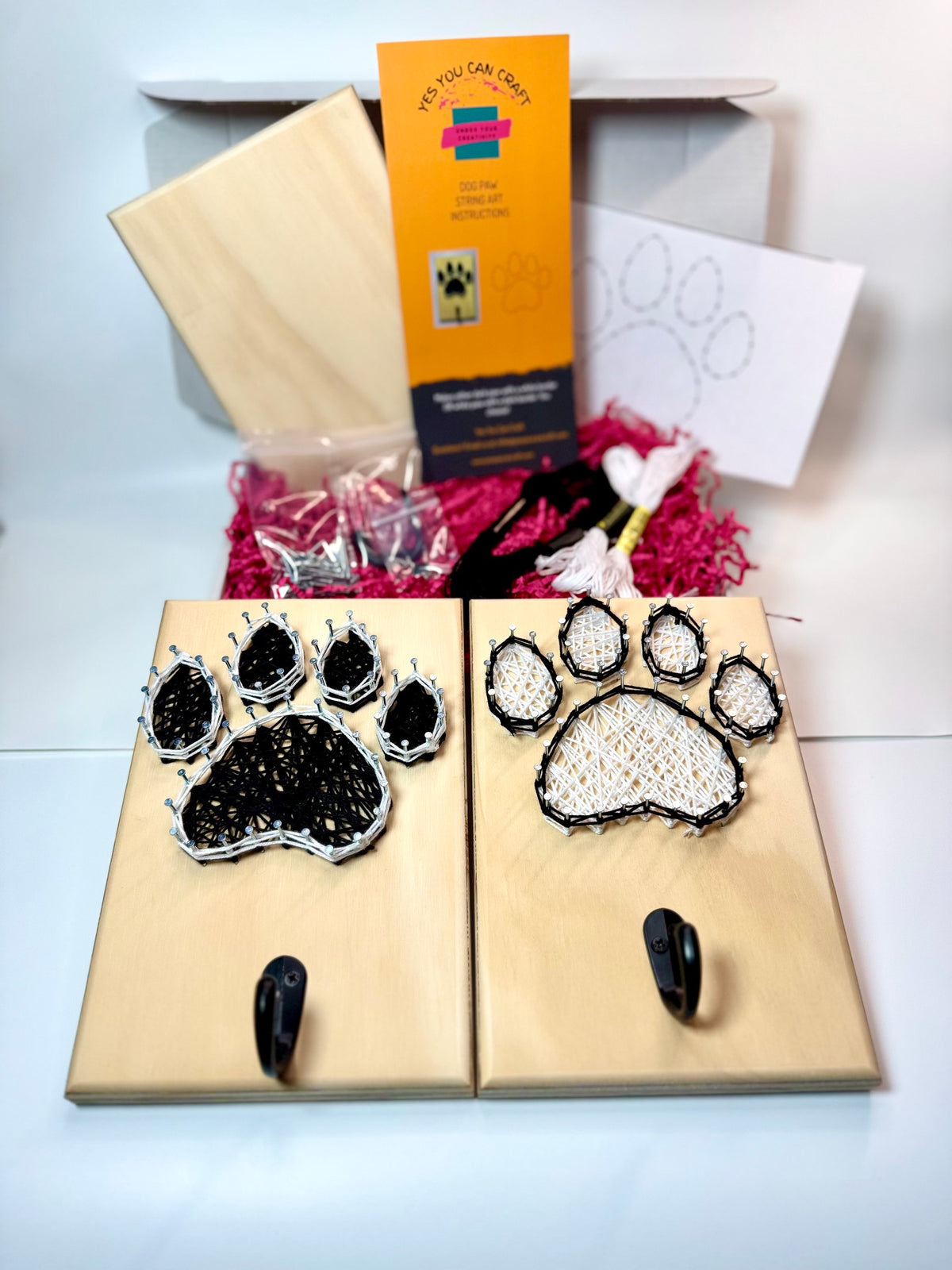 two completed varieties of a dog paw string art with hook. one black and white and the other white with black. DIY craft kit contents in background with board, instructions, nails, hook, thread and template.