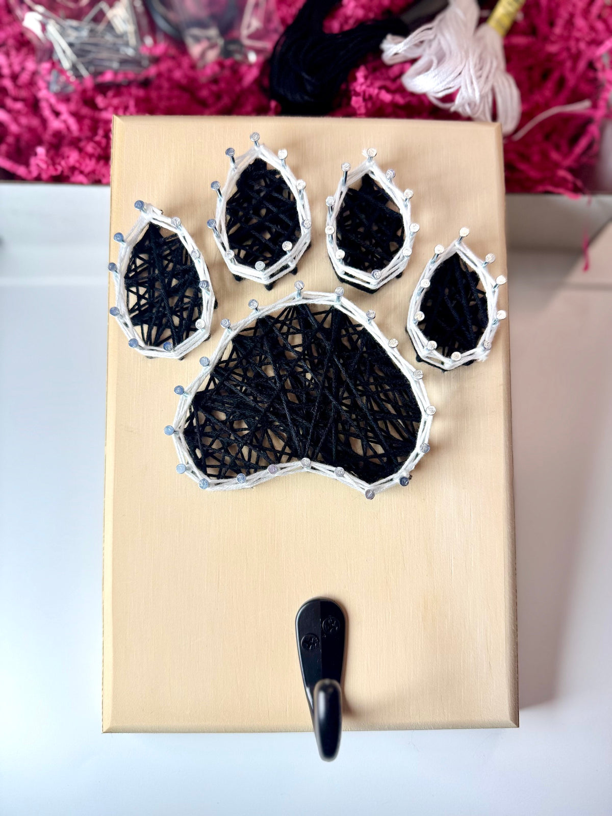 A wooden paw print string art piece with black yarn wrapped around silver nails, mounted on a light wood plaque with a black hook at the bottom, photographed on a white surface with pink paper filler in the background.